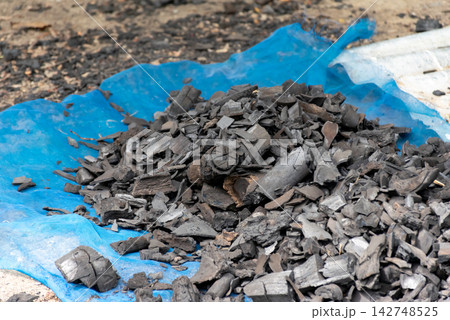 Pile of natural wood charcoal pieces blue mesh sack outdoors representing traditional fuel production biomass energy cooking charcoal industry Pile of natural wood charcoal pieces blue mesh sack outdoors representing traditional fuel production biomass energy cooking charcoal industry 142748525