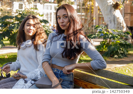Two women sit on a bench in a park while other people walk by on a sunny day in autumn Two women sit on a bench in a park while other people walk by on a sunny day in autumn 142750349