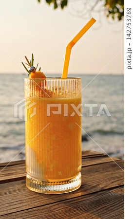 Closeup Of Tropical Drink With Straw On Wooden Table By Sea In Warm Sunlight. Summer Refreshment And Seaside Vacation Concept. Closeup Of Tropical Drink With Straw On Wooden Table By Sea In Warm Sunlight. Summer Refreshment And Seaside Vacation Concept. 142753789