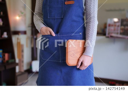Adult Asian cafe owner in blue apron holding wooden tray standing in modern coffee shop interior Adult Asian cafe owner in blue apron holding wooden tray standing in modern coffee shop interior 142754149