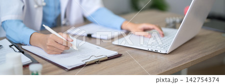 Female doctor writing medical notes on clipboard while colleague types on laptop in modern clinic office Female doctor writing medical notes on clipboard while colleague types on laptop in modern clinic office 142754374