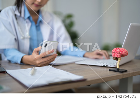 Asian female doctor reviewing patient notes on smartphone at modern clinic desk with brain model and laptop Asian female doctor reviewing patient notes on smartphone at modern clinic desk with brain model and laptop 142754375