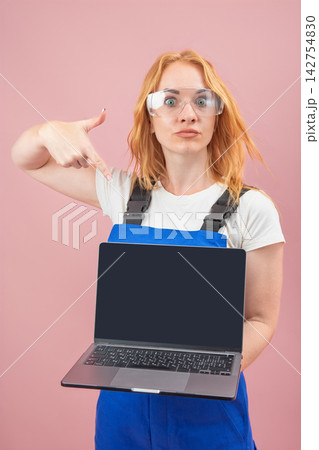 Female professional in work overalls and safety glasses pointing finger at blank laptop screen, communicating an important message or new information on a pink background Female professional in work overalls and safety glasses pointing finger at blank laptop screen, communicating an important message or new information on a pink background 142754830