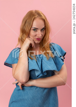 Woman with red hair and green eyes smirking playfully, holding a hand to her chin while posing with a confident and skeptical expression against a pink background Woman with red hair and green eyes smirking playfully, holding a hand to her chin while posing with a confident and skeptical expression against a pink background 142754834