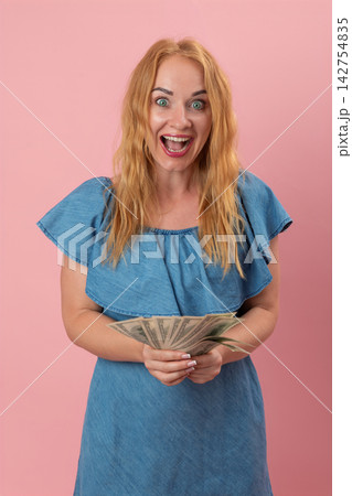 Excited woman with wide eyes and an open mouth celebrating a financial win, holding a fan of United States currency banknotes against a pink background Excited woman with wide eyes and an open mouth celebrating a financial win, holding a fan of United States currency banknotes against a pink background 142754835
