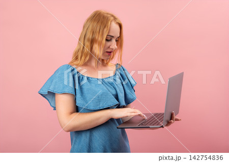 Young woman standing against a pink background, holding an open laptop, and typing on the keyboard while looking at the screen Young woman standing against a pink background, holding an open laptop, and typing on the keyboard while looking at the screen 142754836