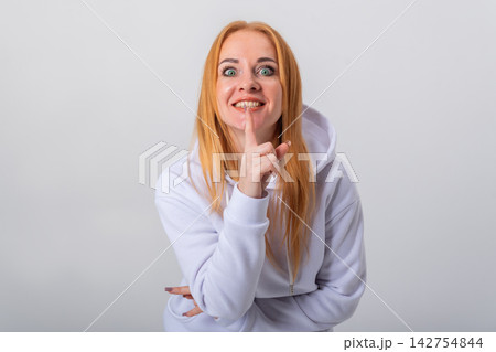 Young woman with red hair and a bright smile pressing her index finger to her lips, conveying a powerful message of silence and secrecy with wide eyes and an expressive face Young woman with red hair and a bright smile pressing her index finger to her lips, conveying a powerful message of silence and secrecy with wide eyes and an expressive face 142754844