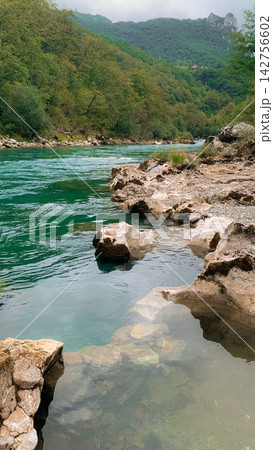 Turquoise river water flowing alongside rugged rocky riverbanks in a lush mountain valley landscape featuring deep green trees and overcast soft daylight during a serene summer afternoon retreat 142756602