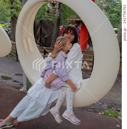 Mother embracing daughter inside circular playground seat with ropes course, trees, and wooden fencing behind them at outdoor adventure park, showing family affection, care, and leisure 142756761