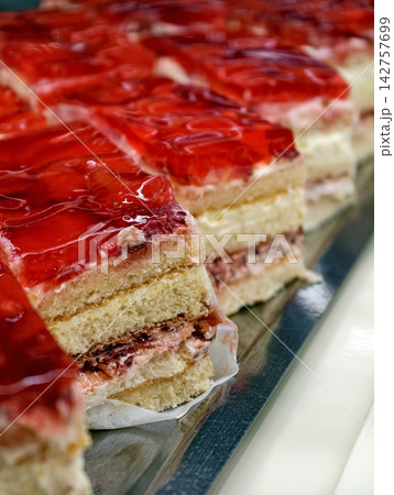 Layered pastries with creamy filling and red jelly on top presented on a metal tray, creating an inviting display of sweet desserts in a pastry shop showcase Layered pastries with creamy filling and red jelly on top presented on a metal tray, creating an inviting display of sweet desserts in a pastry shop showcase 142757699