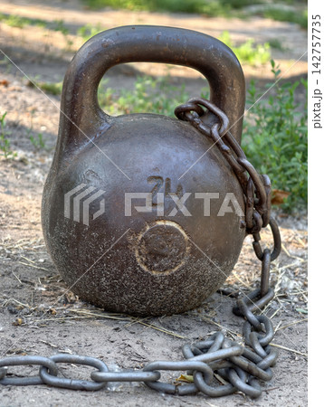 Old iron 24 kg kettlebell standing outdoors on the ground with a metal chain at a sports area for weight training and physical culture exercises Old iron 24 kg kettlebell standing outdoors on the ground with a metal chain at a sports area for weight training and physical culture exercises 142757735