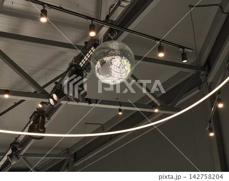 Disco ball chandelier hangs from industrial ceiling with spotlights, track lighting, and suspended mirrored beams in modern showroom interior with glamorous reflective sparkle Disco ball chandelier hangs from industrial ceiling with spotlights, track lighting, and suspended mirrored beams in modern showroom interior with glamorous reflective sparkle 142758204