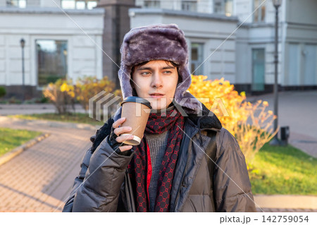 Androgynous man in fur hat drinking takeaway coffee outdoors. Young stylish male walking in city park during autumn. Concept of modern urban lifestyle and daily routine in cold weather. Androgynous man in fur hat drinking takeaway coffee outdoors. Young stylish male walking in city park during autumn. Concept of modern urban lifestyle and daily routine in cold weather. 142759054