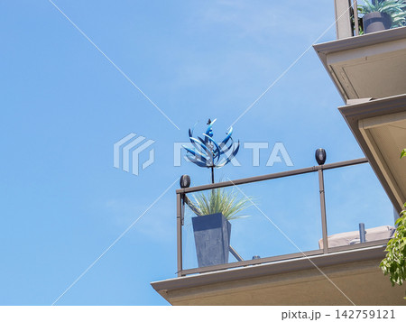 Blue decorative metal wind spinner on a glass-railed modern apartment balcony against a clear blue sky - June 13, 2026 Ruston WA, USA Blue decorative metal wind spinner on a glass-railed modern apartment balcony against a clear blue sky - June 13, 2026 Ruston WA, USA 142759121