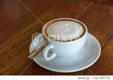 Hot cappuccino coffee topped creamy milk foam served white ceramic cup saucer spoon warm wooden table cozy cafe setting Hot cappuccino coffee topped creamy milk foam served white ceramic cup saucer spoon warm wooden table cozy cafe setting 142763759