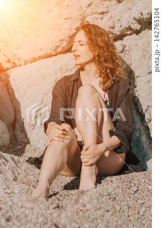 Woman ocean beach sitting relaxing near rocky cliff in sunlit nature enjoying a peaceful summer vacation atmosphere 142765104