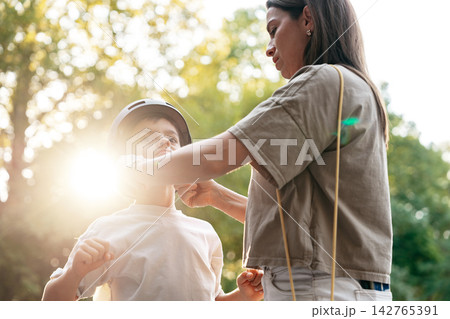 Mother helps son adjust helmet while preparing for outdoor activity in park during daytime Mother helps son adjust helmet while preparing for outdoor activity in park during daytime 142765391