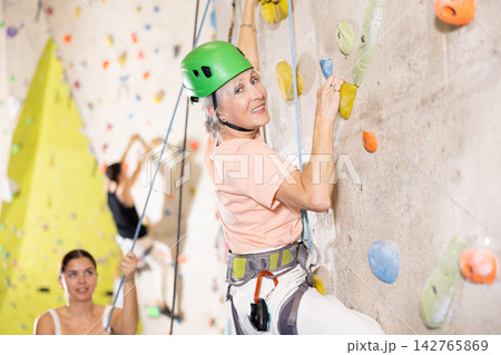 positive muscular retired woman in gym climbing up artificial rock climbing wall 142765869