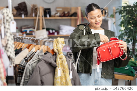 Asian female customer standing against background of shelves with clothes in modern store and choosing handbag 142766420