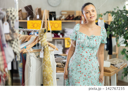 Asian woman chooses sundress in store and carefully examines her reflection in mirror Asian woman chooses sundress in store and carefully examines her reflection in mirror 142767235