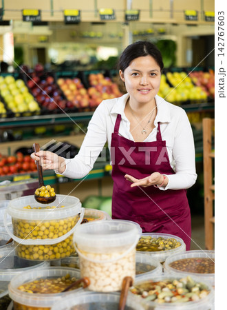Positive asian girl seller in apron offers pickled olives in supermarket Positive asian girl seller in apron offers pickled olives in supermarket 142767603