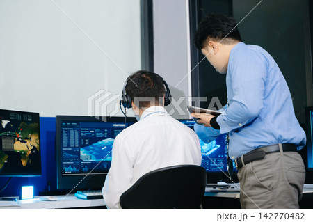 Team IT operator in a headset monitors global logistics and facility data across multiple screens. A modern network control room focused on digital surveillance Team IT operator in a headset monitors global logistics and facility data across multiple screens. A modern network control room focused on digital surveillance 142770482