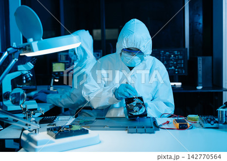 Cleanroom technicians in protective suits repairing a computer hard drive. Engineers conducting digital forensics and data recovery Cleanroom technicians in protective suits repairing a computer hard drive. Engineers conducting digital forensics and data recovery 142770564