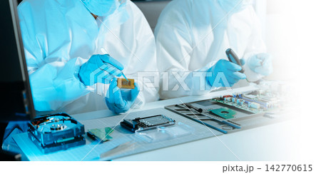 Cleanroom technicians in protective suits examining microchips and motherboards with a magnifying glass. Engineers conducting precise electronic repairs Cleanroom technicians in protective suits examining microchips and motherboards with a magnifying glass. Engineers conducting precise electronic repairs 142770615