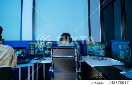 Military officers in camouflage monitor global tactical data and radar on multiple screens within a high tech cybersecurity command center. Military officers in camouflage monitor global tactical data and radar on multiple screens within a high tech cybersecurity command center. 142770617