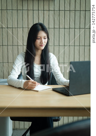 Gen Z Young Woman Studying with Laptop and Notebook at Wooden Desk in Modern Interior Gen Z Young Woman Studying with Laptop and Notebook at Wooden Desk in Modern Interior 142775754
