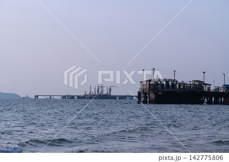Distant pier stands silent against calm sea. Sky holds soft haze, blending with horizon Distant pier stands silent against calm sea. Sky holds soft haze, blending with horizon 142775806