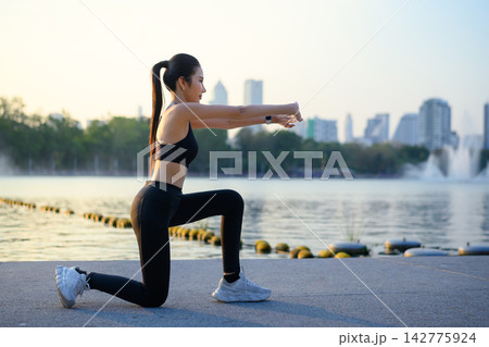 Woman raises arms stretching by lake with city view at sunrise Woman raises arms stretching by lake with city view at sunrise 142775924
