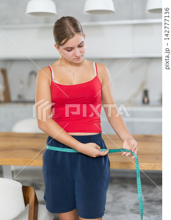 Teenage girl measures her waist and chest with measuring tape in kitchen 142777136