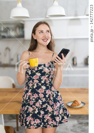 Woman in dress standing in kitchen, looking at mobile phone and drinking coffee 142777223