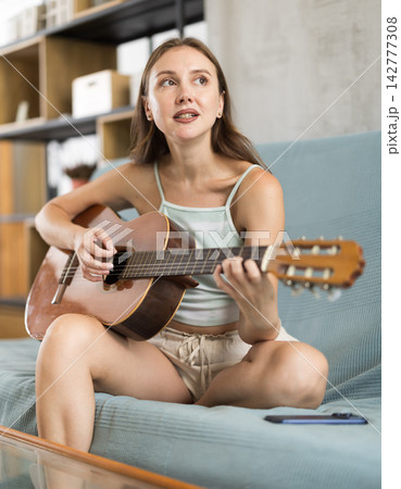 Woman practicing guitar in living room sitting on gray sofa Woman practicing guitar in living room sitting on gray sofa 142777308