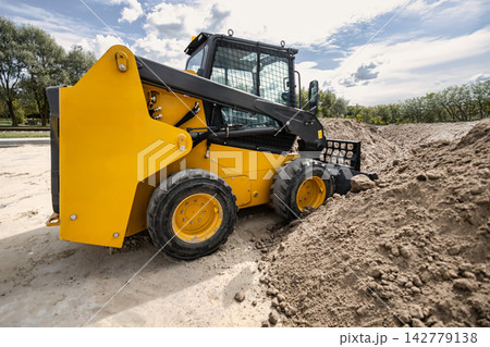 A mini loader is working on a landscaping site, moving earth and gravel to prepare for project completion. It is parked near a pile of dirt A mini loader is working on a landscaping site, moving earth and gravel to prepare for project completion. It is parked near a pile of dirt 142779138