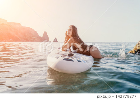 Woman Paddleboard Ocean Relaxing - A woman lays on a paddleboard in the ocean, looking out at the rocky cliffs in the distance. Woman Paddleboard Ocean Relaxing - A woman lays on a paddleboard in the ocean, looking out at the rocky cliffs in the distance. 142779277