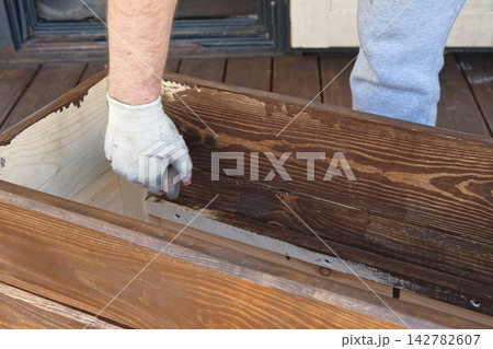 Hand in glove painting wooden box with brown oil stain brush. Worker applying protective varnish on pine timber planks surface, DIY woodwork. Wood staining process. Backyard Woodworking, Maintenance. Hand in glove painting wooden box with brown oil stain brush. Worker applying protective varnish on pine timber planks surface, DIY woodwork. Wood staining process. Backyard Woodworking, Maintenance. 142782607