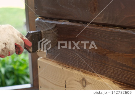 Hand in glove painting wooden box with brown oil stain brush. Worker applying protective varnish on pine timber planks surface, DIY woodwork. Wood staining process. Backyard Woodworking, Maintenance. Hand in glove painting wooden box with brown oil stain brush. Worker applying protective varnish on pine timber planks surface, DIY woodwork. Wood staining process. Backyard Woodworking, Maintenance. 142782609