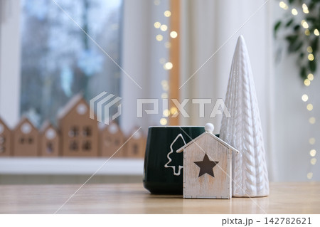 Green ceramic mug with fir tree contour stands on wooden table. Festive winter holiday scene with small wooden house ornament and ceramic cone object against blurred decorated Christmas backdrop. 142782621