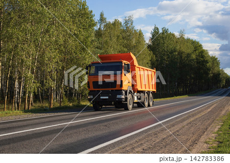 Orange dump truck on a forest road. Construction equipment driving on the highway. Orange dump truck on a forest road. Construction equipment driving on the highway. 142783386