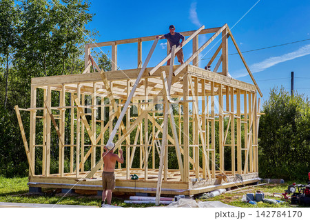 Rafter system installation, young worker positions timber rafters on frame house while second builder assists with roof structure construction. Rafter system installation, young worker positions timber rafters on frame house while second builder assists with roof structure construction. 142784170