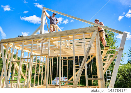 Ridge purlin mounted, young workers position rafters above it at construction site. 142784174