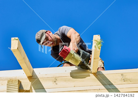 Pneumatic framing nailer drives nails into wooden boards while young worker performs carpentry during timber frame construction. 142784192