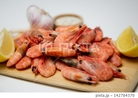 A close-up side view of frozen shrimps on a wooden board, two lemon slices diagonally, a salt shaker and a head of garlic next to it. Concept: an expanded set of seafood seasonings. High quality photo A close-up side view of frozen shrimps on a wooden board, two lemon slices diagonally, a salt shaker and a head of garlic next to it. Concept: an expanded set of seafood seasonings. High quality photo 142784370