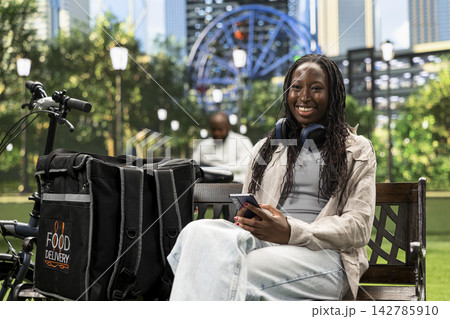 Portrait of smiling courier looking at the camera seated with smartphone, checking on demand mobile app for new order. Delivery person with thermal backpack on bench. Portrait of smiling courier looking at the camera seated with smartphone, checking on demand mobile app for new order. Delivery person with thermal backpack on bench. 142785910
