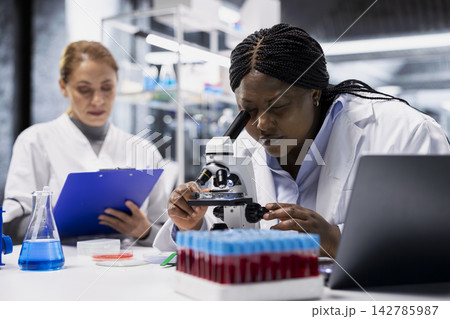 Female scientists examining bacterial cultures on slide using lab microscope. Team of laboratory specialist ensuring consumer safety by checking for microscopic pathogens with optical device Female scientists examining bacterial cultures on slide using lab microscope. Team of laboratory specialist ensuring consumer safety by checking for microscopic pathogens with optical device 142785987
