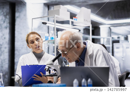 Senior scientist inspecting new drug compound under microscope, helped by assistant reporting findings. Elderly man in research facility verifying medication quality, intern apprentice taking notes Senior scientist inspecting new drug compound under microscope, helped by assistant reporting findings. Elderly man in research facility verifying medication quality, intern apprentice taking notes 142785997