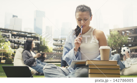 Focused student writing in notebook beside open book, taking notes and studying alone on campus lawn. Young woman does study session with stacked books and backpack. Focused student writing in notebook beside open book, taking notes and studying alone on campus lawn. Young woman does study session with stacked books and backpack. 142786005