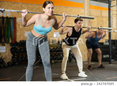 Energetic young female athlete practicing sit-ups with rod near other people gym Energetic young female athlete practicing sit-ups with rod near other people gym 142787095